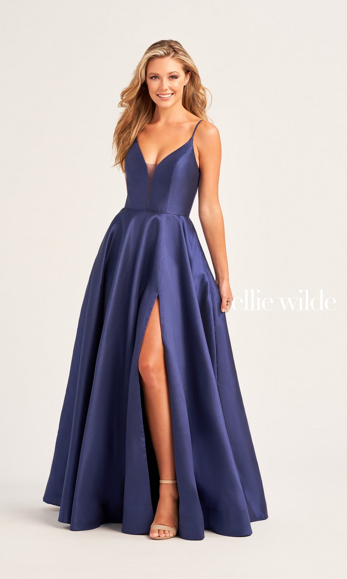 A woman in the Ellie Wilde Long A-Line Satin Prom Dress EW35232, featuring a sleeveless design, strappy back, and thigh-high slit, stands smiling against a plain light background.